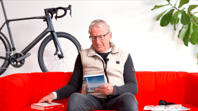 Ein Mann sitzt auf einem roten Sofa und liest ein Buch. Im Hintergrund steht ein Fahrrad und eine Zimmerpflanze.