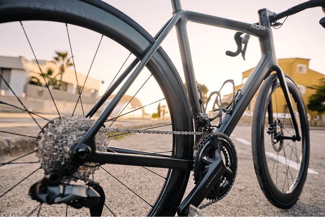 Close-up of a sleek black road bike with visible gearset and chain, parked on a street at sunset.